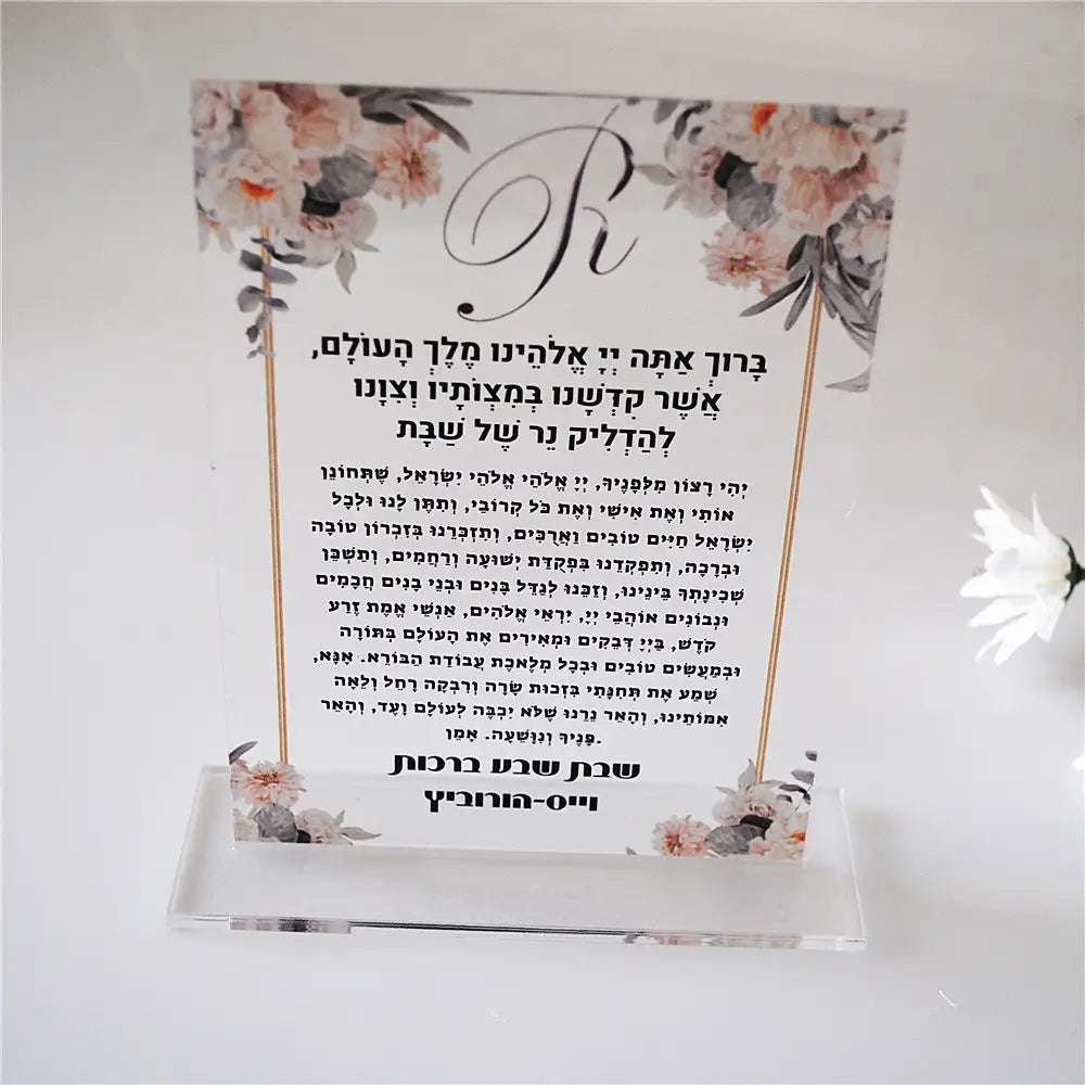 A clear acrylic wedding table sign with a floral design of delicate pink and white flowers framed by gold-toned borders and Hebrew text, part of a custom acrylic Shabbat candle blessing souvenir set of 10 pieces.