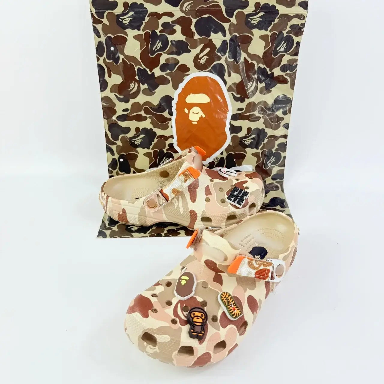 Beige and brown camouflage-patterned bape x crocs clogs with orange straps and ape head-themed charms designed as anti slip beach shoes for model toys gifts.