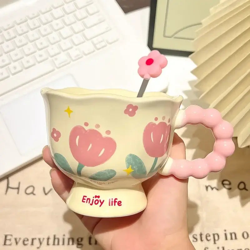 Une tasse tulipe en céramique crème pastel avec des décors de fleurs roses, une anse rose perlée et la mention « enjoy life » en rouge, en écriture manuscrite, sur sa base, dans une boîte cadeau à motif floral.