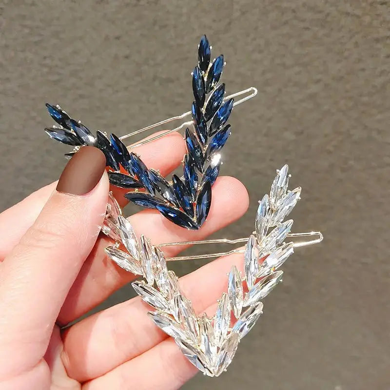 A crystal v shape hair clip set featuring a pair of elegant silver-toned metal hair clips with leaf designs, one adorned with deep blue crystals and the other with clear crystals.
