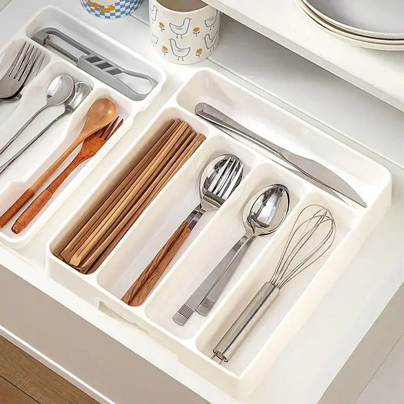 Organisateur de tiroir à couverts en plastique blanc, avec des compartiments segmentés contenant des cuillères en bois, des fourchettes métalliques, des couteaux et un fouet pour le rangement des ustensiles de cuisine.