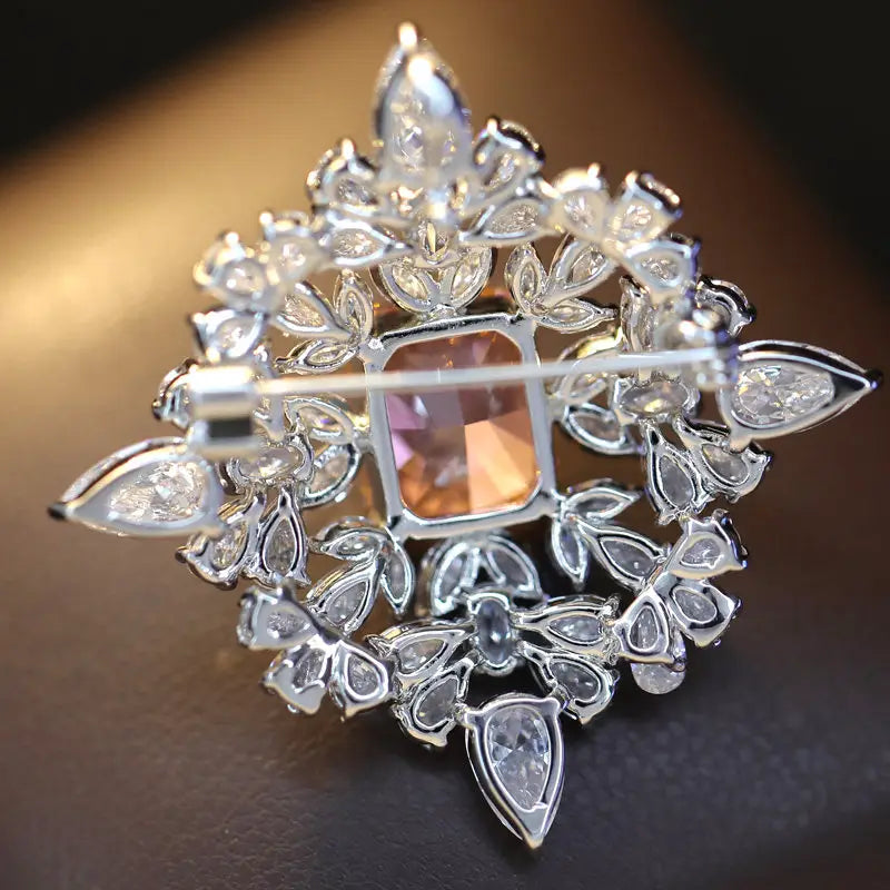 Pin de lujo de diseñador en forma de broche de mariposa de zirconio, con una base plateada brillante, una gema rectangular central en tono rosa melocotón, rodeada de diseños intrincados de hojas y pétalos, adornados con cristales facetados transparentes.