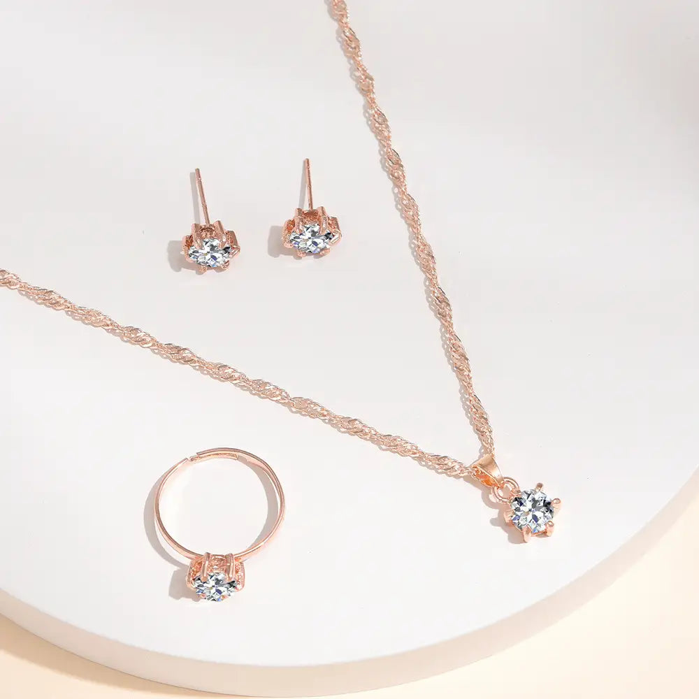 Juego de joyería de 5 piezas con circonitas cúbicas de color dorado rosado, con un collar de cadena trenzada que presenta un dije central con apariencia de diamante, aretes tipo botón a juego y un anillo ajustable adornado con piedras de cristal transparentes.