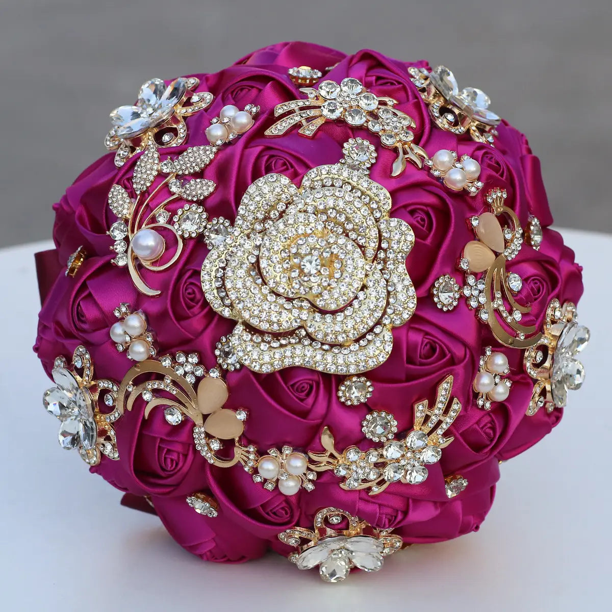 Un bouquet de mariée vibrant en satin fuchsia décoré de broches florales aux tons dorés, de strass étincelants et d’accents de perles.