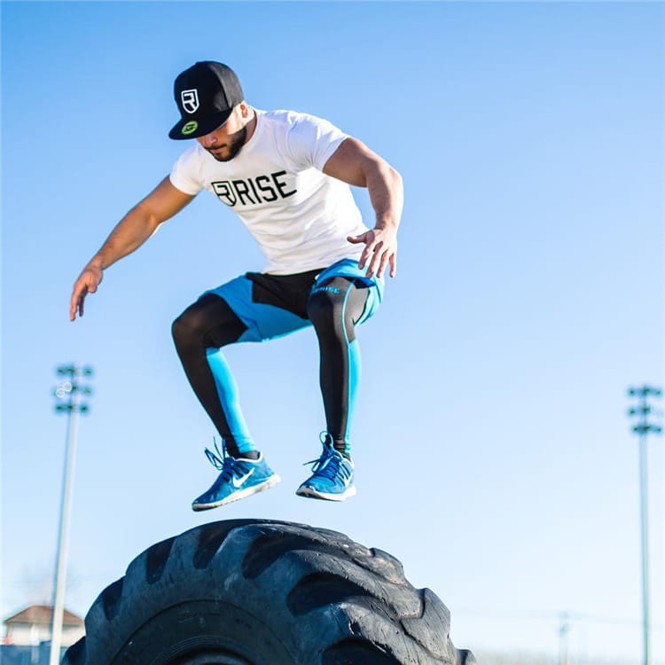 Ein Mann, der ein weißes Herren-Workout-T-Shirt mit Gold-Rise-Logo und schwarzem Rise-Aufdruck trägt, springt über einen großen schwarzen Gummireifen.