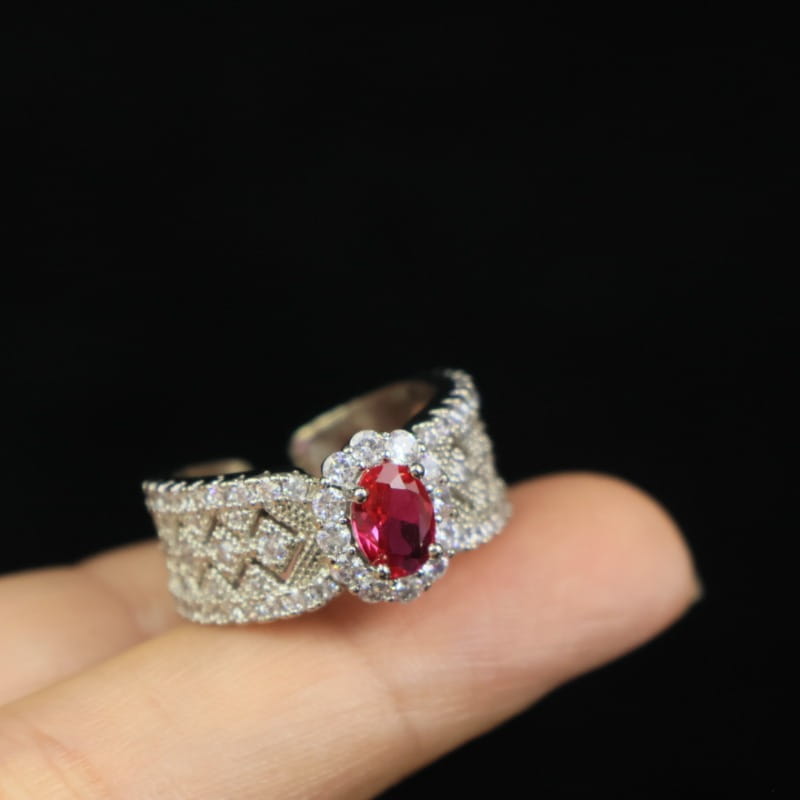 Ein Silberband-Rubin-Halo-Ring mit einem markanten ovalen roten Rubin, umgeben von kleineren klaren Diamanten und einem kunstvollen Diamant-Pavé-Muster entlang der Seiten.