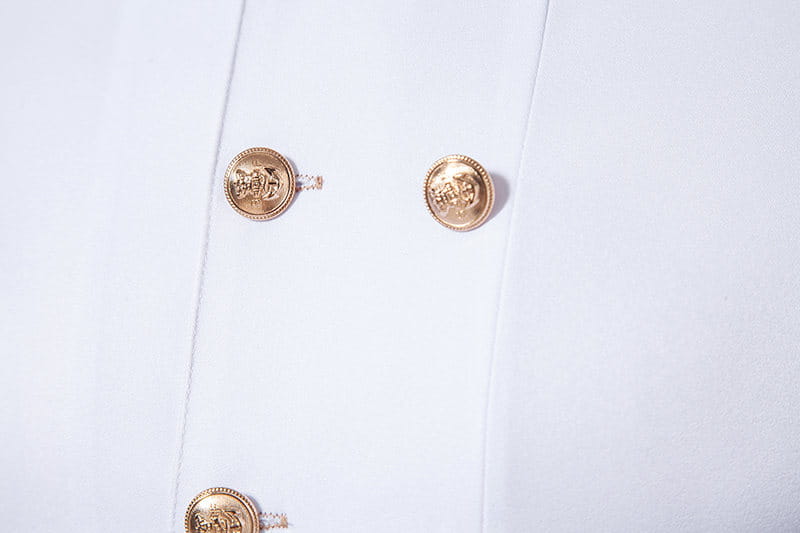 Botones circulares de tono dorado con logotipos en relieve y bordes decorados con cuentas adornan una camisa gótica blanca para hombre con cuello alzado y cruzada doble.