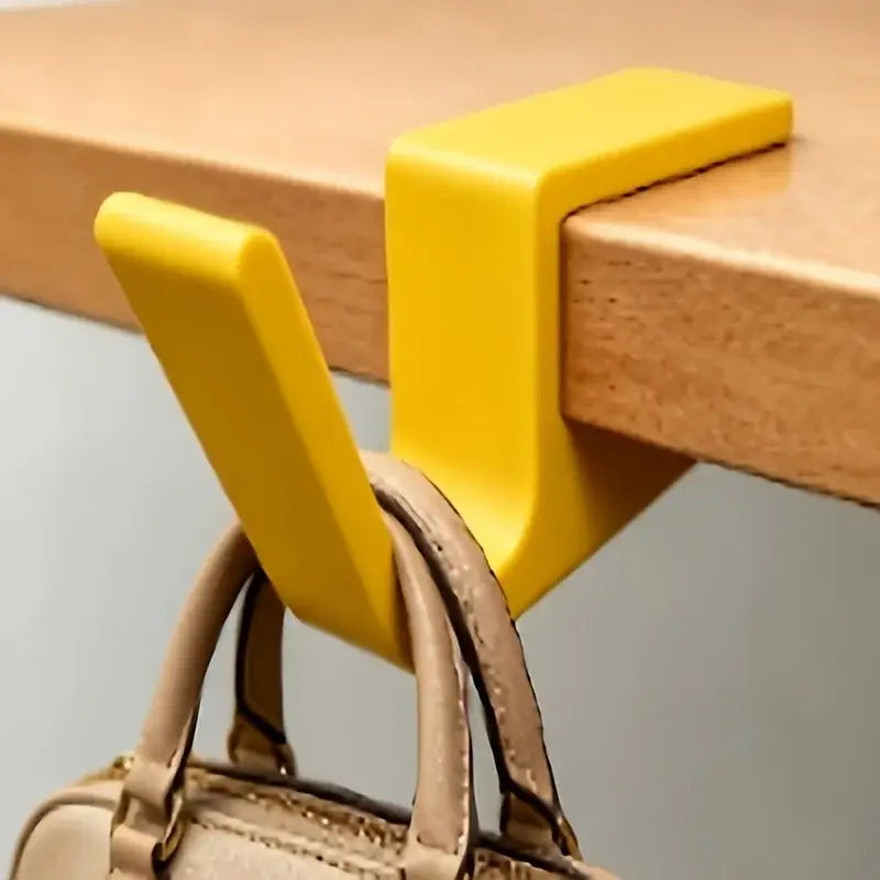 Bright yellow plastic desk edge bag hook mounted on a desk holding a beige handbag strap.