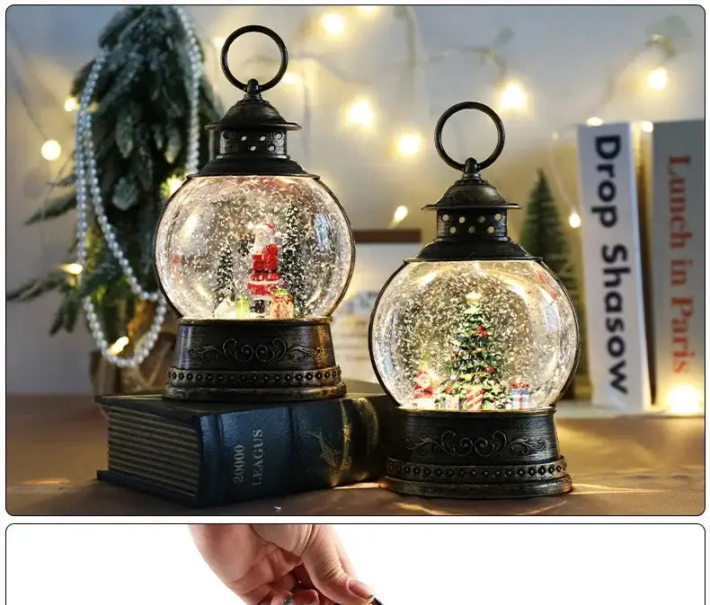 Two black lantern-style christmas snow globe music boxes with clear glass spheres containing miniature christmas scenes and falling glitter resting on decorative book bases.