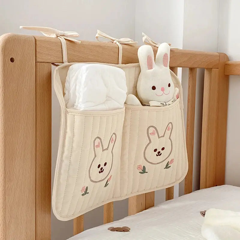 クマの刺繍が施されたクリーム色のキルティング製ベビーカー用オーガナイザー袋（布製）で、刺繍のうさぎの顔、花柄のディテール、そしてポケットが2つ付いており、オムツとぬいぐるみのおもちゃを収納できます。
