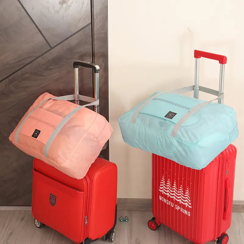 Juego de bolsa de lona de viaje plegable con bolsas plegables rosas y celeste claro con detalles de cremallera grises, apoyadas sobre maletas rojas.
