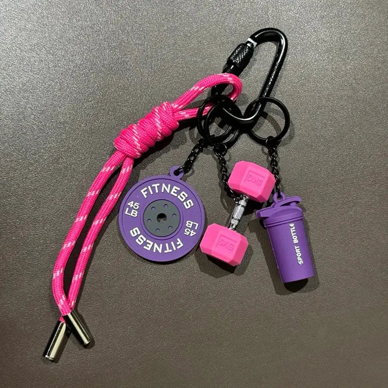 Un porte-clés de musculation vibrant rose et violet comprenant un cordon tressé avec une breloque de disque de poids circulaire gravée Fitness 45 lb, deux breloques de mini-haltères et une petite bouteille shaker, pour les amateurs de fitness.