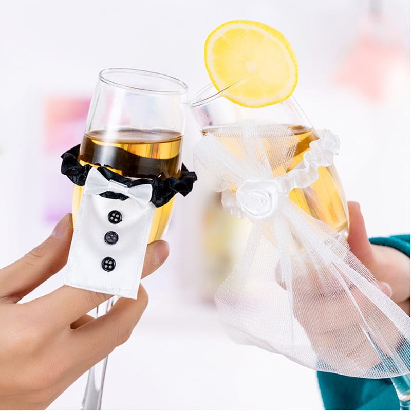 Dos copas tipo champán con envolturas decorativas de tela con temática de esmoquin y vestido de novia en un juego de cubiertas de copas de vino para la novia y el novio.