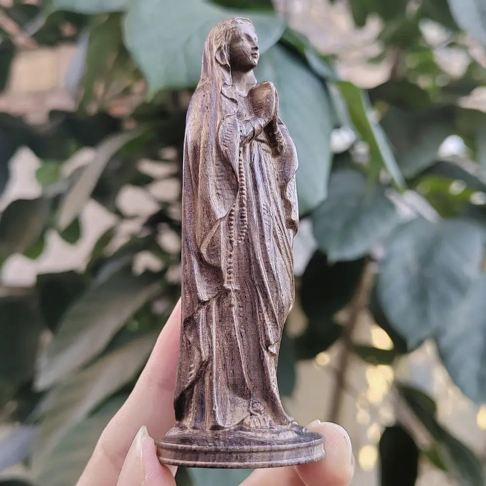 Statuette de la Vierge Marie en bois sculpté à la main, représentant une figure vêtue d’une robe détaillée tenant un chapelet, avec des sculptures complexes et une finition brun chaud.