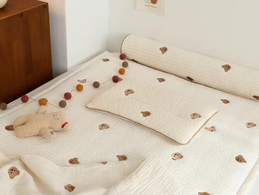 散りばめられた茶色のクマのアプリケをあしらい、柔らかな質感のある生地が特徴のクリーム色のキルティング刺繍コットンのベビーピロー。