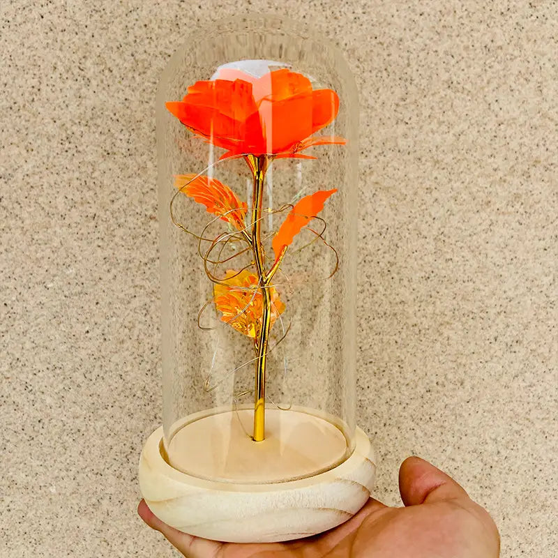 Eine kräftig orangefarbene, goldfolierte „Eternal Gold Rose“ mit metallischem Stiel und Blattwerk aus Draht, ausgestellt in einer klaren Glaskuppel auf einem hellfarbigen Marmorsockel, mit LED-Licht-Geschenk-Funktionen.