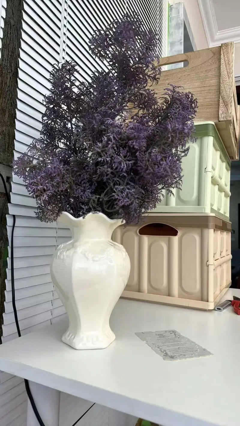 Un jarrón de cerámica moderno hecho de cerámica blanca con borde acanalado y detalles florales en relieve que sostiene un arreglo exuberante de follaje artificial morado intenso, perfecto para la decoración del hogar con estilo de cerámica moderna francesa.