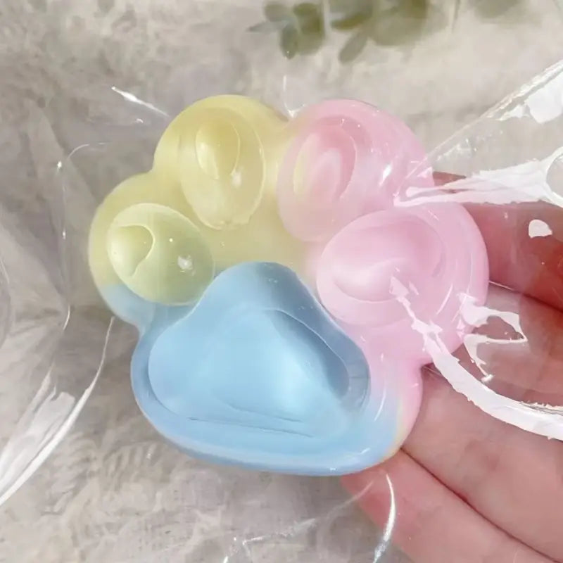 Dispositif anti-stress en silicone pastel et translucide, en forme de patte, avec un dégradé doux du jaune au rose puis au bleu, présenté comme un jouet anti-stress souple et moelleux en griffe de chat arc-en-ciel.