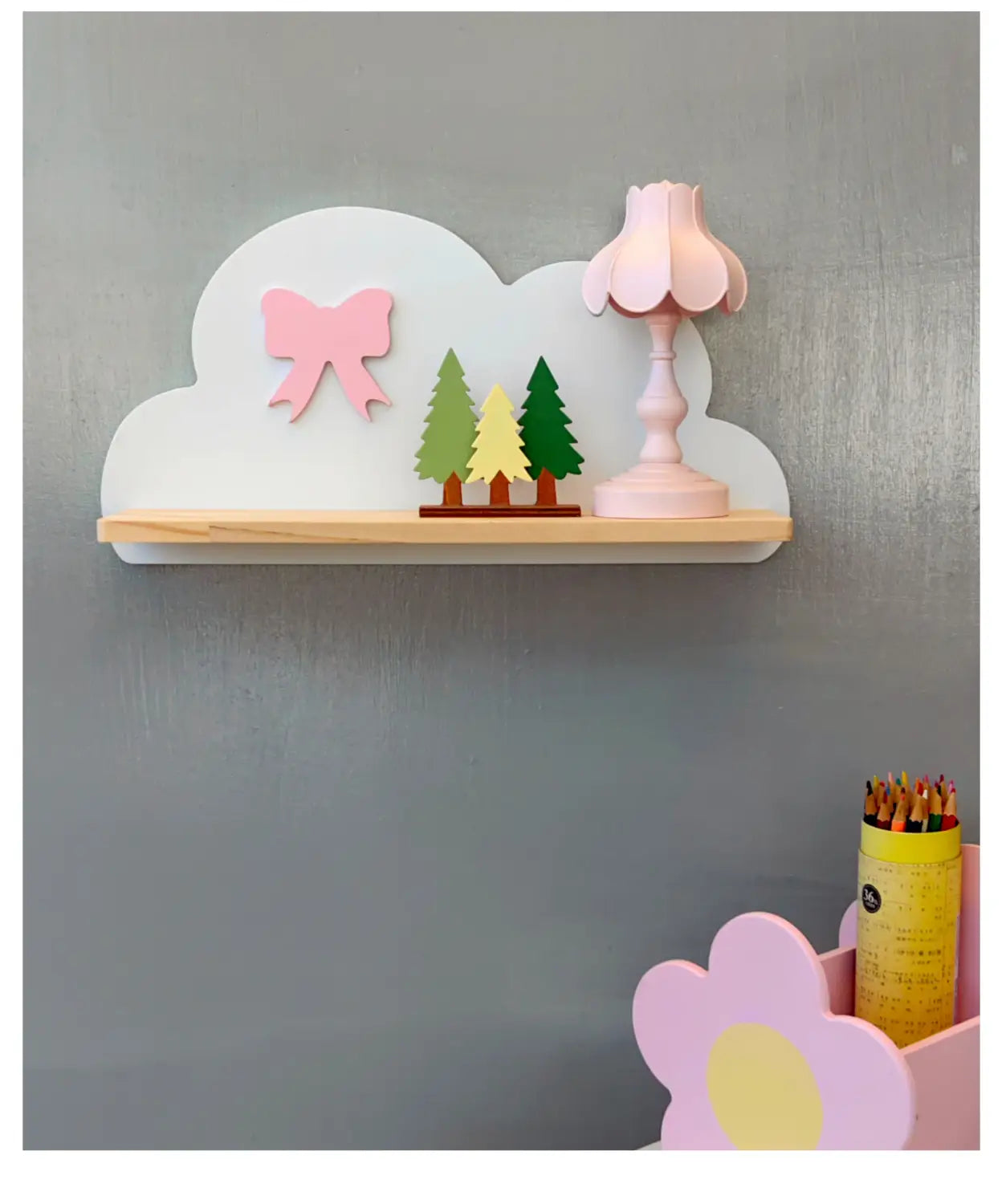 Ein pastelrosa und weißes, wolkenförmiges Bücherregal aus Holz für Kinder an der Wand mit einem Holzsockel verfügt über eine dekorative Schleife, drei Miniaturbäume in Grün und Gelb sowie eine passende rosa Lampe.
