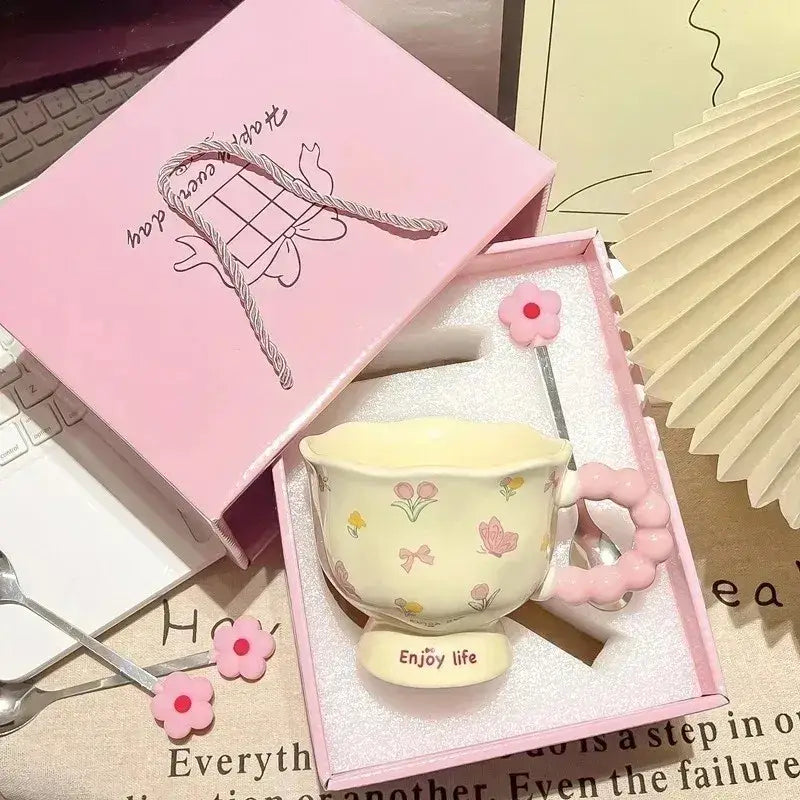 Tasse tulipe en céramique jaune pastel avec des décors floraux et de papillons roses, une anse rose perlée texturée et la mention « enjoy life » sur sa base, dans une boîte cadeau à motif floral.