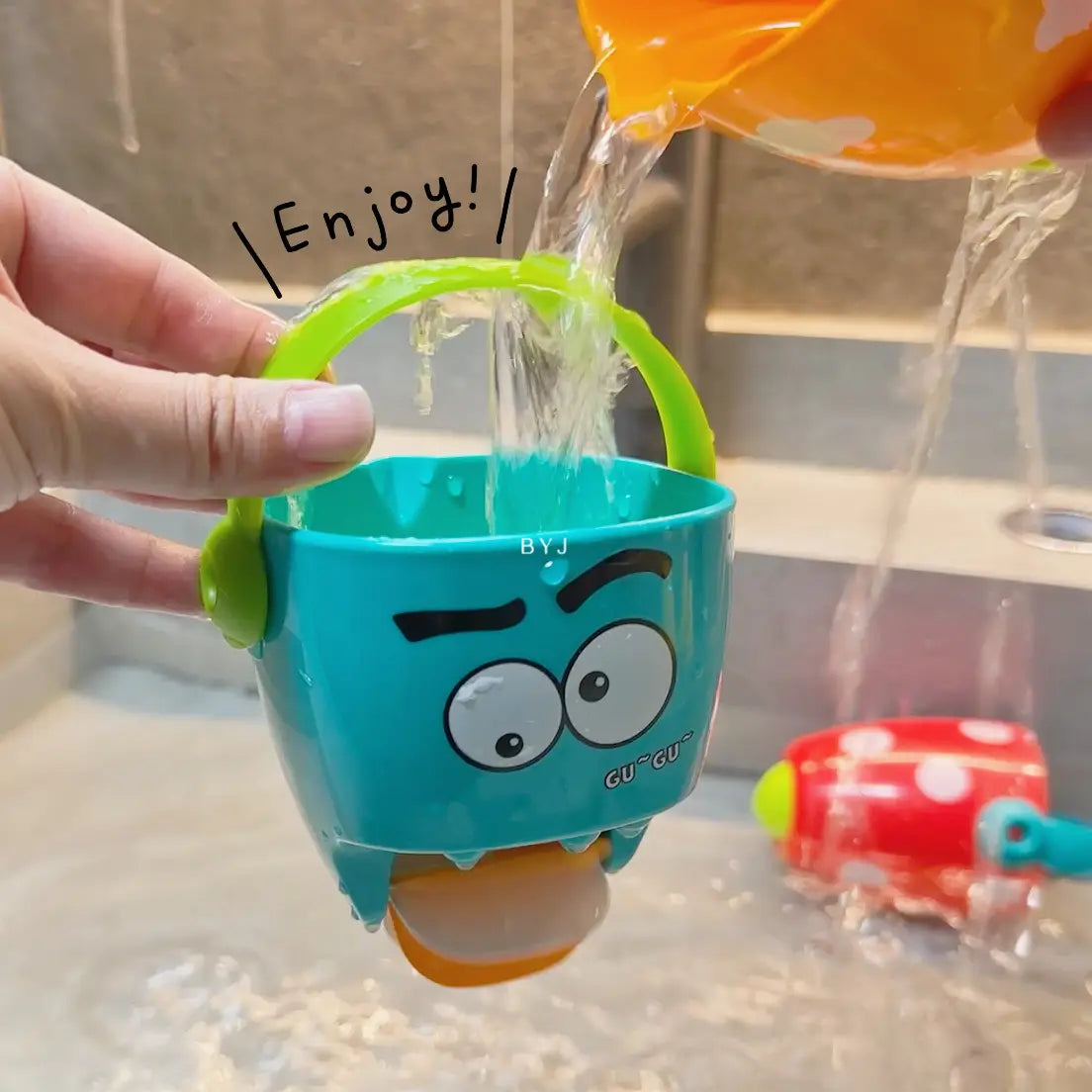 Ein türkisfarbener Kunststoff-Badewannen-Eimer für Babys mit grünem Griff und einem Design mit Cartoon-Monster-Gesicht aus dem Spielset für Baby-Badewannen-Eimer mit Wassersprühfunktion.