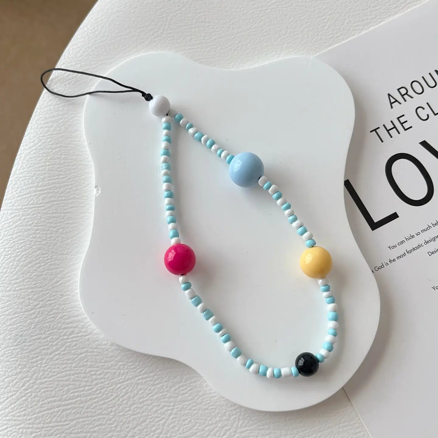 un bracelet de téléphone à perles colorées avec des perles sphériques bleu clair, blanches, roses, jaunes et noires enfilées sur un cordon fin.