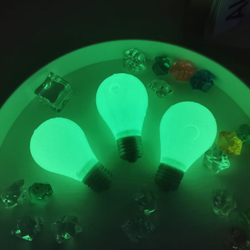 Tres juguetes antiestrés tipo gomita con forma de bombilla de luz verde luminosa, con bases negras en rosca, dispuestos en un patrón circular sobre una superficie blanca, rodeados por cubitos de hielo de plástico transparente y de colores.