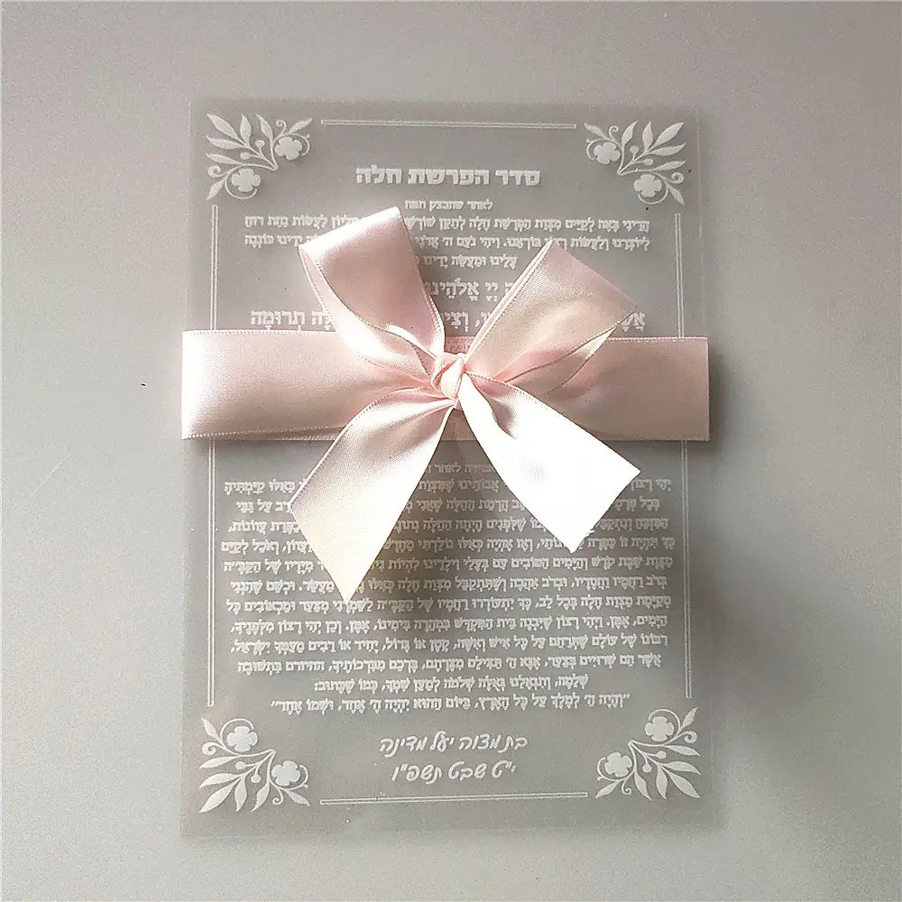 A delicate translucent acrylic prayer card with elegant silver floral borders and Hebrew text tied with a soft pink satin ribbon bow for custom Hebrew acrylic prayer card wedding and Bar Mitzvah souvenir.