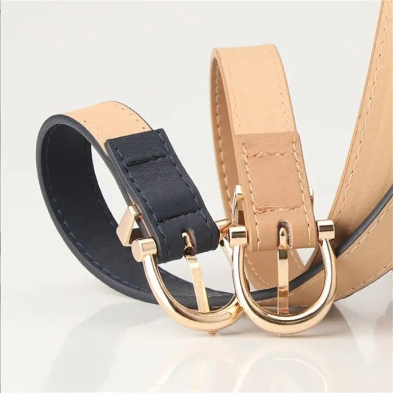 Dos cinturones delgados de piel sintética (PU) en azul marino y beige con hebillas tipo galgo (horsebit) de tono dorado para las cinturas de mujer.