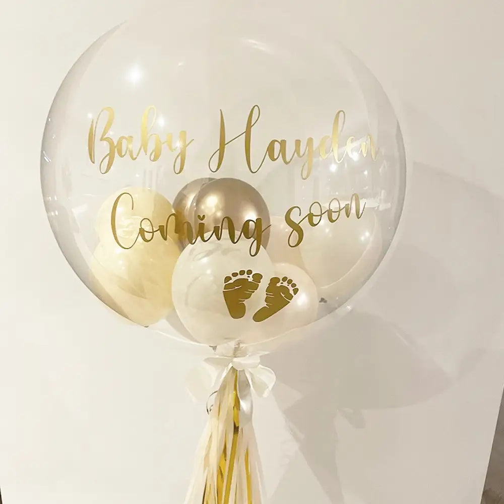 Individueller Baby-Party-Ballonaufkleber auf einem transparenten 36-Zoll-Bobo-Ballon, mit goldfarbener Folienbeschriftung „Baby Hayden coming soon“ sowie kleinen weißen Ballons im Inneren, darunter einer mit goldfarbenen Fußabdrücken.