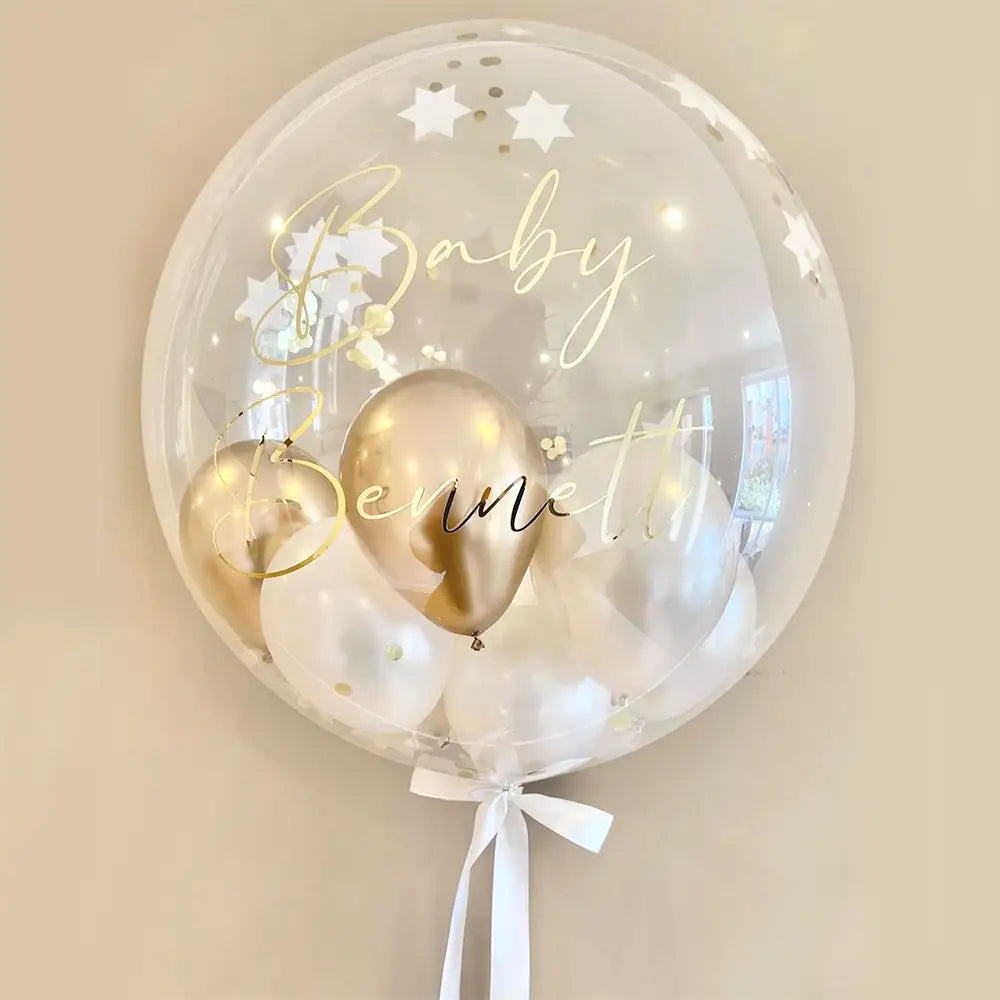 Individueller Baby-Party-Ballonaufkleber auf einem transparenten 36-Zoll-Bobo-Ballon mit goldfarbener geschwungener Schrift „Baby Bennett“ und weißem Stern-Konfetti, enthaltend kleinere metallische goldene sowie transparente Ballons, gebunden mit einer zarten weißen Schleife.