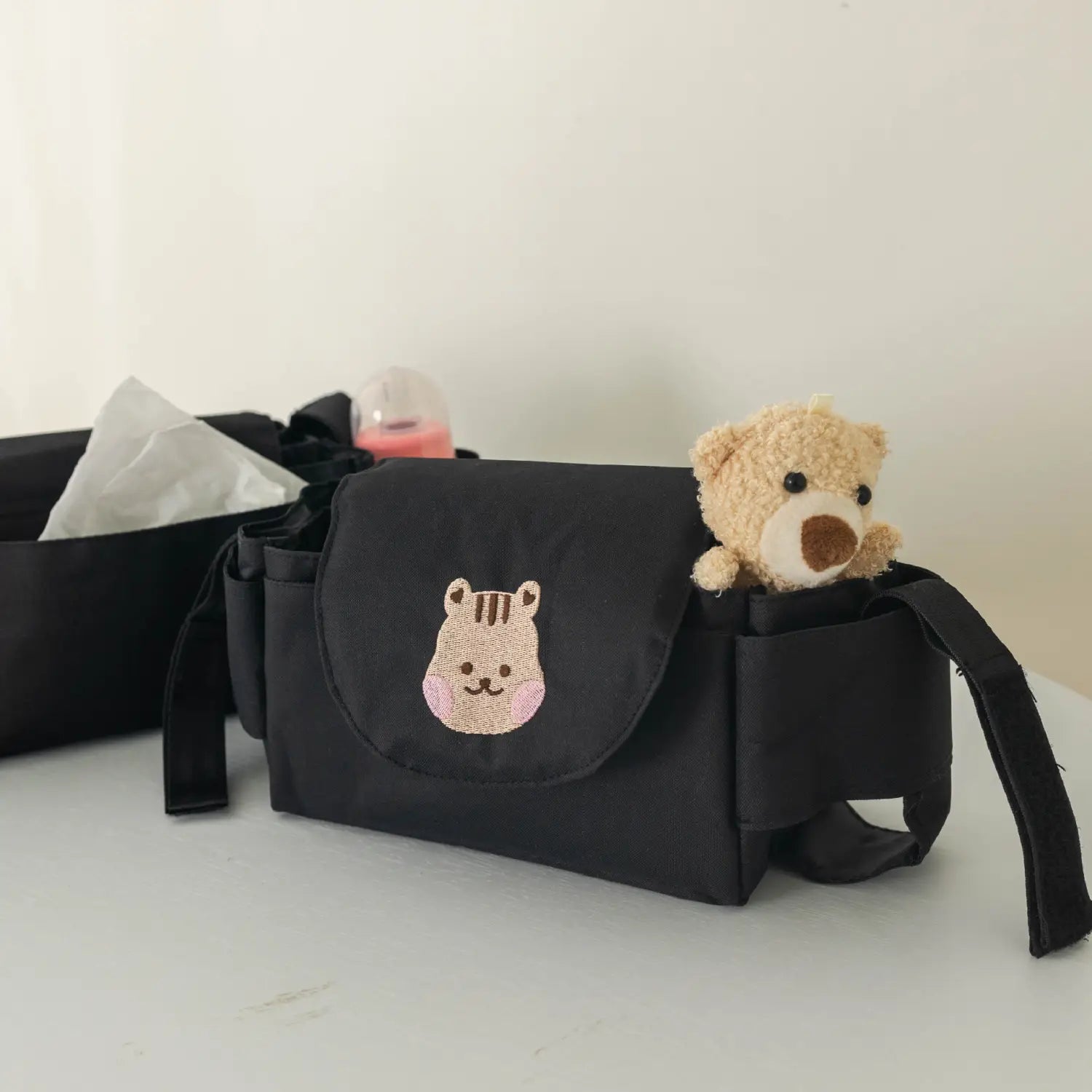 ベビーシート背面に取り付けるサイドポケットからぬいぐるみのテディベアがのぞく、刺繍のトラの顔パッチ付きブラックの布製ベビーカー用オーガナイザーバッグ。