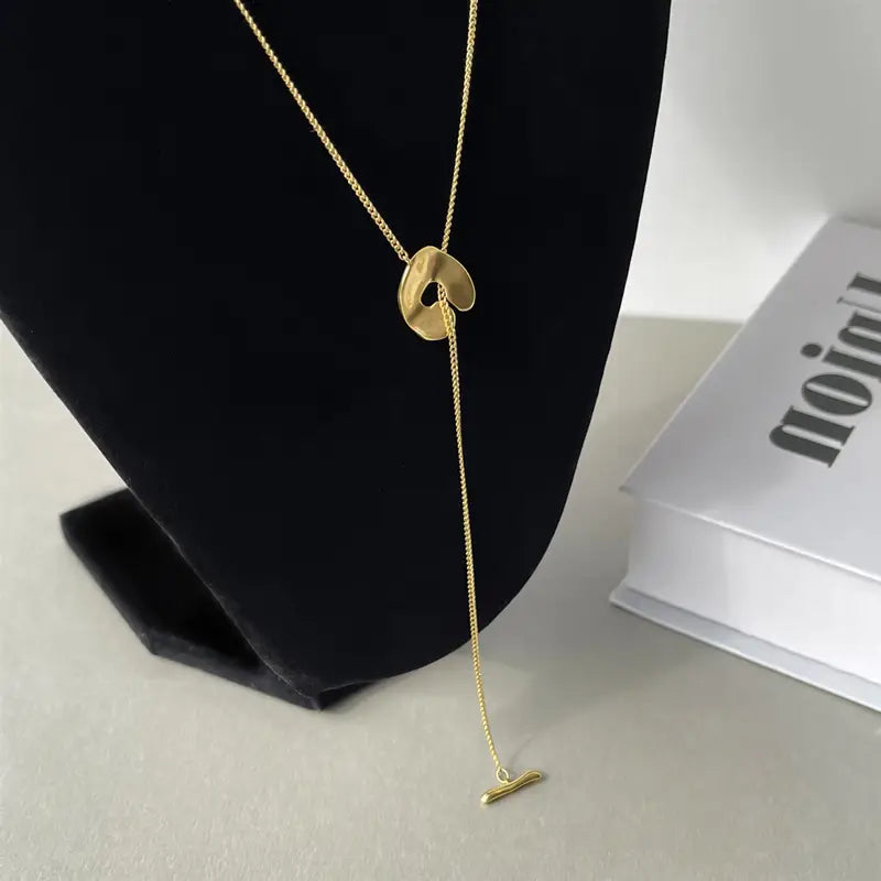 Collier pendentif à superposition géométrique doré pour femmes, présentant un design lariat aux tons dorés avec un pendentif circulaire abstrait épuré et minimaliste ainsi qu'un petit charm en forme de barre.