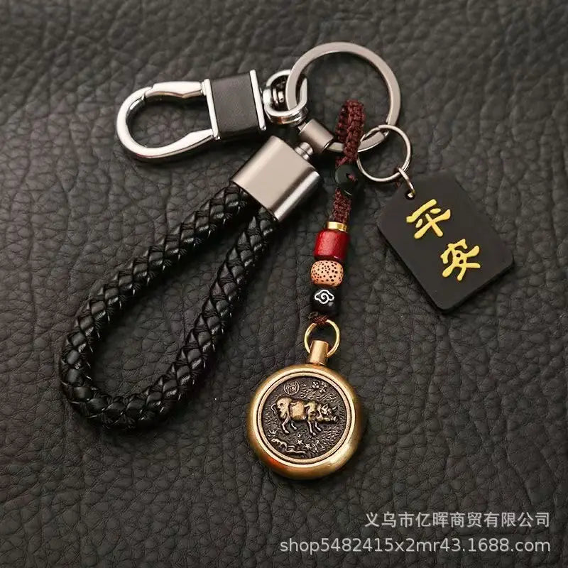 赤と黒のビーズでアクセントを施し、刻印された「牛」の意匠をあしらったブロンズの円形ペンダントを備えた黒の編みレザー製キーチェーン。中国の干支をモチーフにした編みレザー製ラッキーペンダント用の、赤と黒のビーズのほか、小さな長方形のタグ付き。
