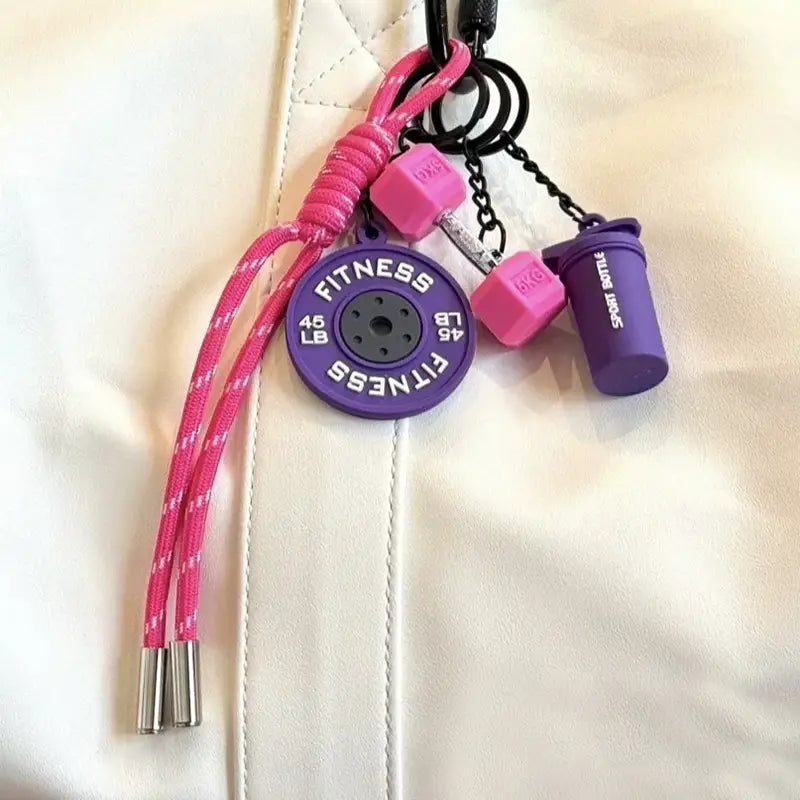 Une breloque pour sac de sport en porte-clés de fitness violet et rose avec un disque de poids circulaire, des breloques d’haltère et une breloque de bouteille shaker.