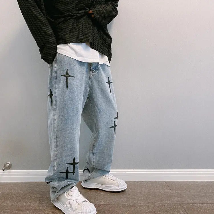 Pantalon cargo en jean à jambes larges à effet délavé clair, avec broderie croisée noire sur les jambes.