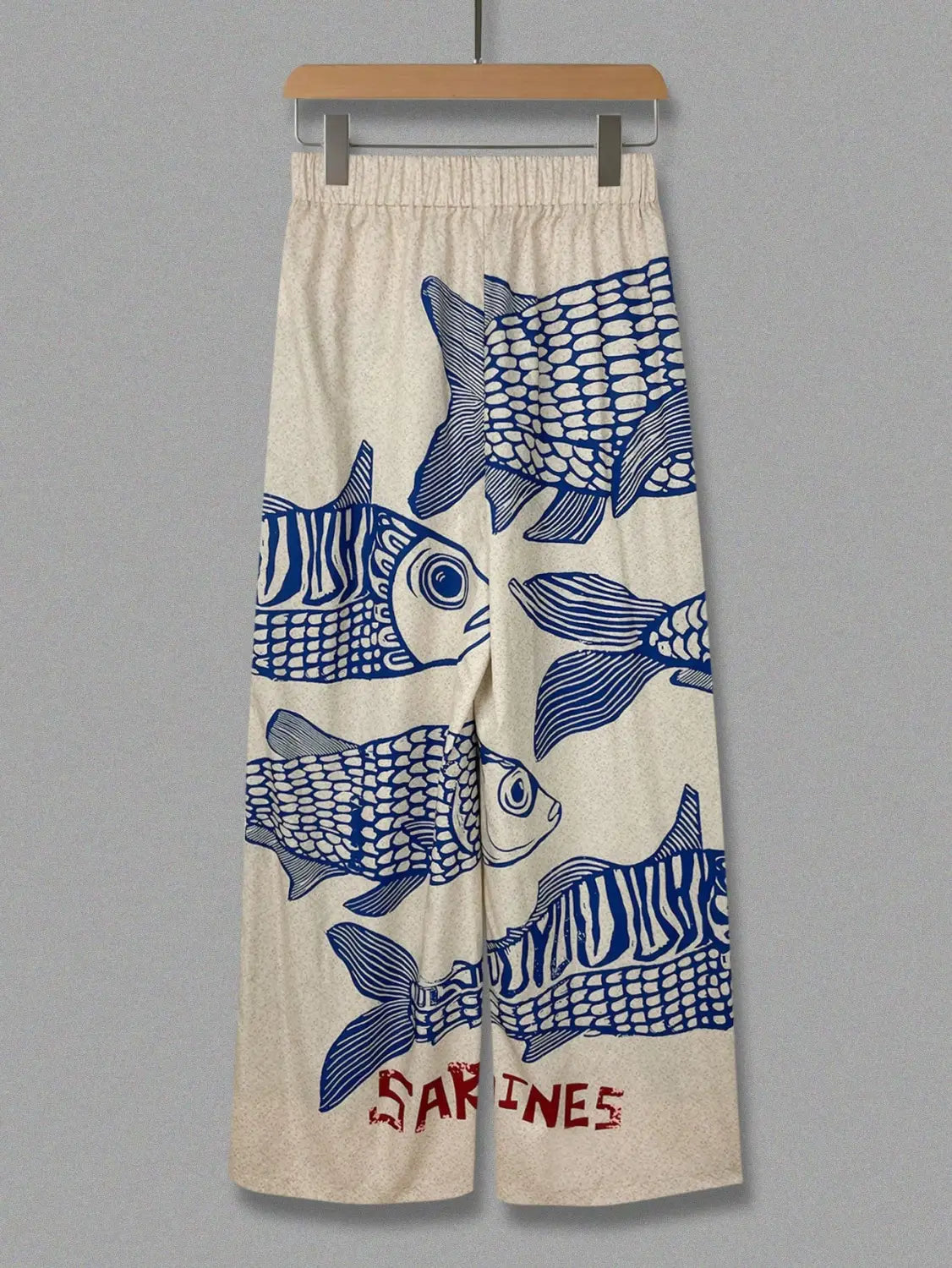 Cream-colored wide leg pants with an elastic waist featuring a bold blue fish print and red sardines lettering at the bottom.