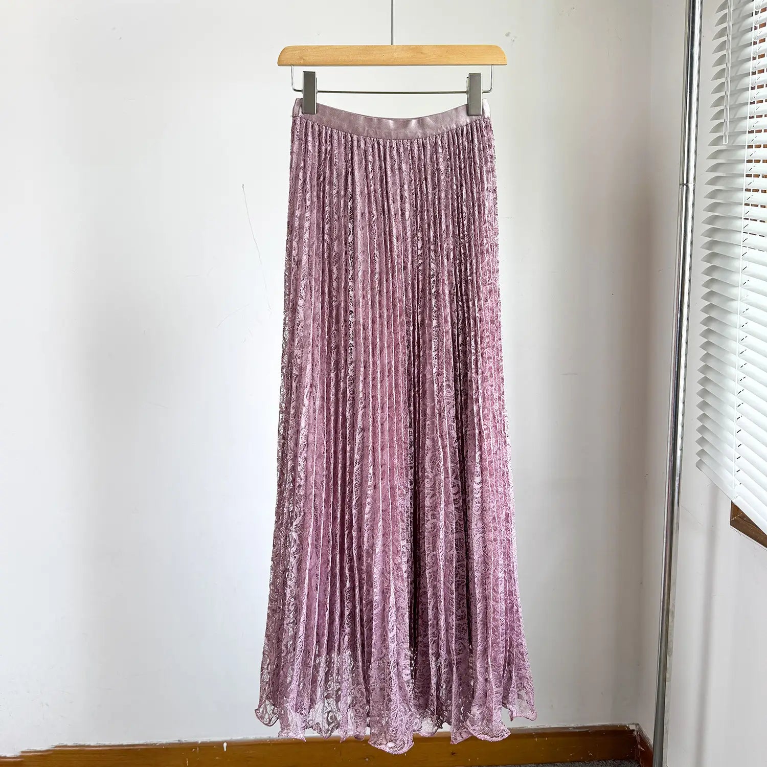 A flowing lavender high waist pleated midi skirt with a subtle lace pattern and satin-like finish hanging on a wooden hanger for y2k streetwear.