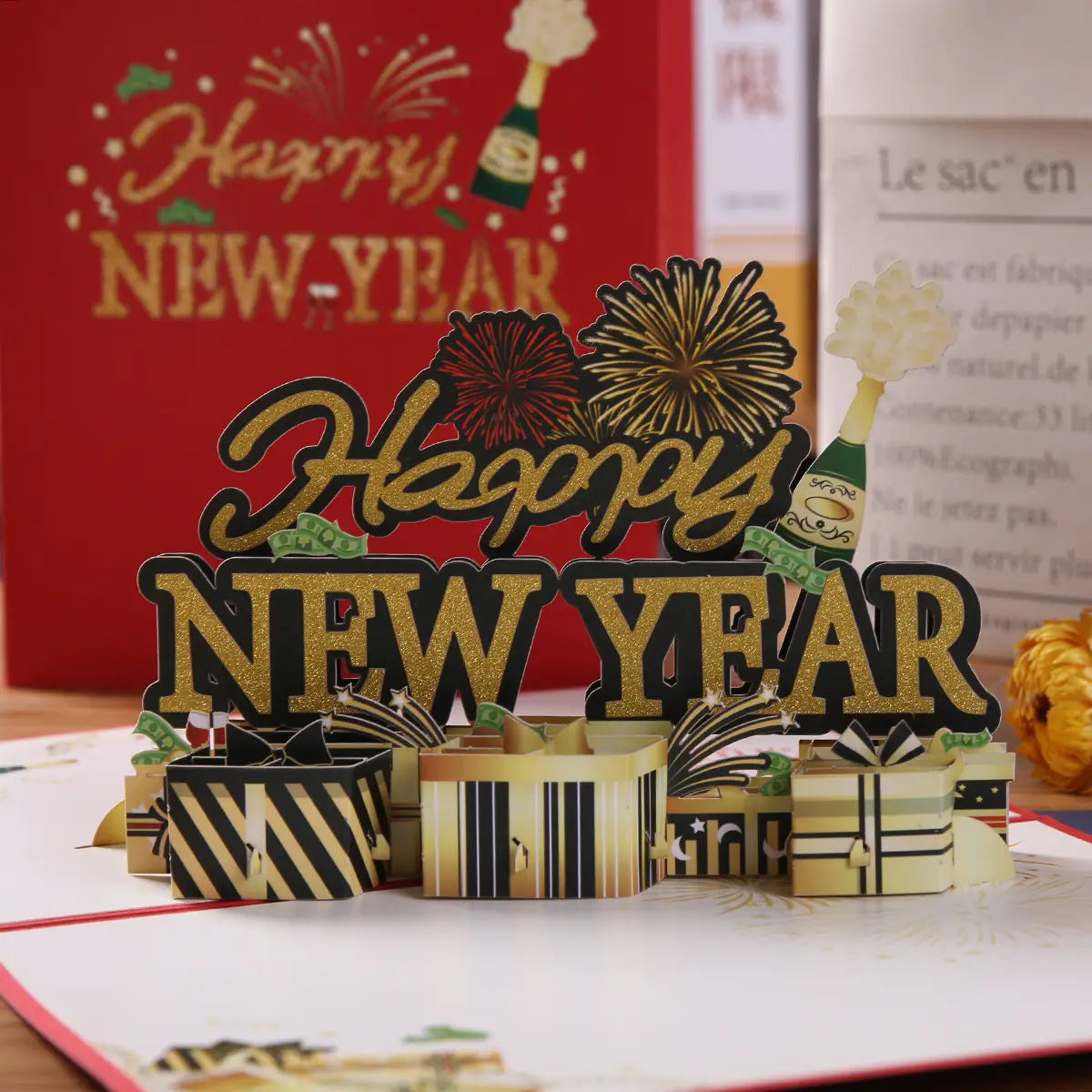 金色のきらめく文字を備えた3Dポップアップ「新年おめでとう」カードで、黒と金のギフトボックス、シャンパンボトル、花火が含まれ、祝祭のための華やかな演出となっております。