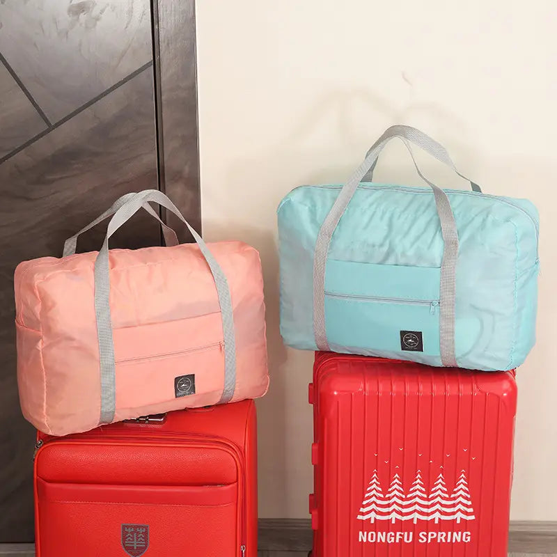 Dos bolsas de lona de viaje plegables ligeras de color pastel con correas grises y parches negros de la marca descansan sobre maletas rojas en un conjunto de bolsa de lona de viaje plegable.