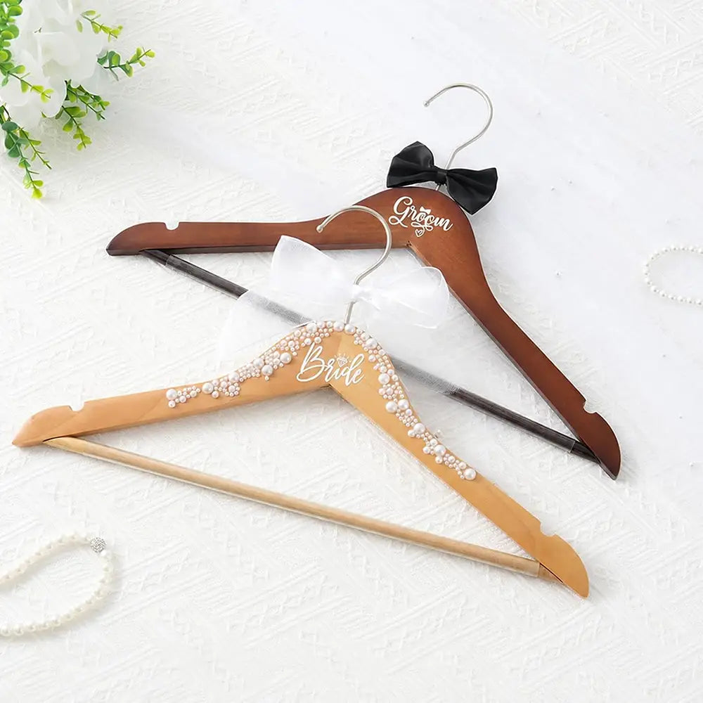 Ensemble de cintres de mariage en bois comprenant un cintre pour la mariée avec une écriture cursive blanche et des ornements perlés, ainsi qu’un cintre pour le marié gravé avec un nœud papillon noir, destiné à l’organisation de la tenue de mariage.