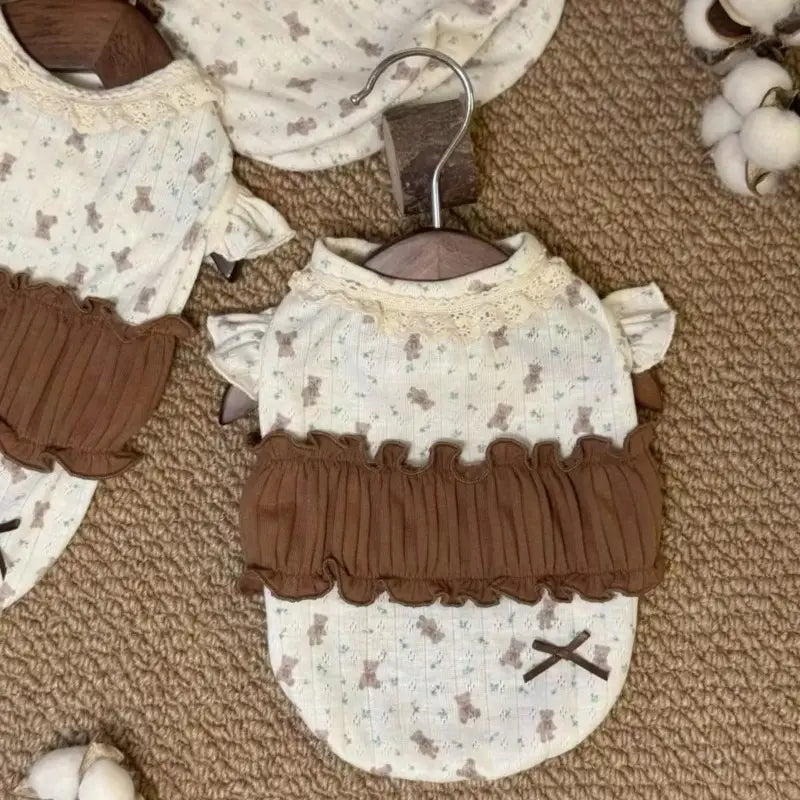 控えめなテディベアのプリントが施された、クリーム色の繊細なフローラルレース犬用ドレスで、ウエスト部分にはブラウンのリブ素材のフリル、首元にはレースのトリムが施されています。