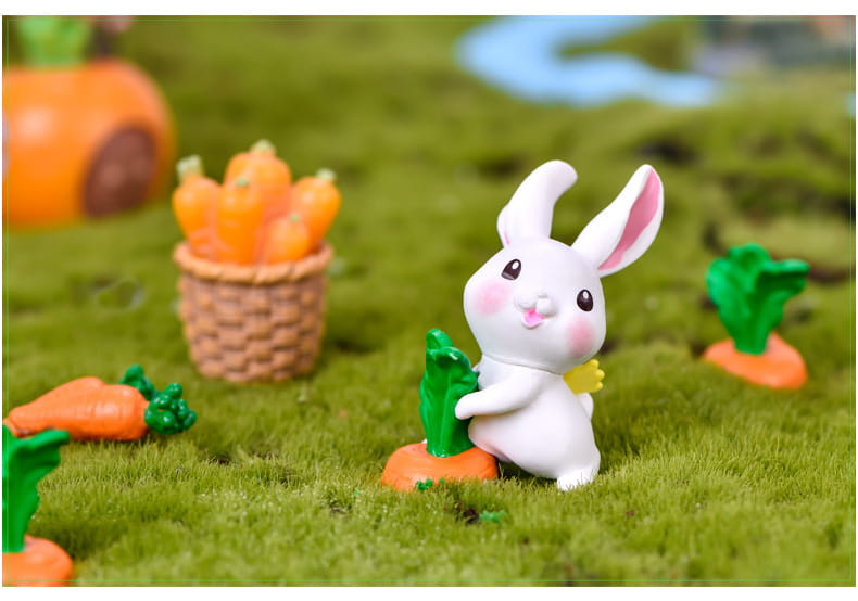 Une figurine miniature en résine blanche de lapin, avec des joues roses et des oreilles internes roses, tenant une carotte verte pour la décoration d’un jardin féerique.