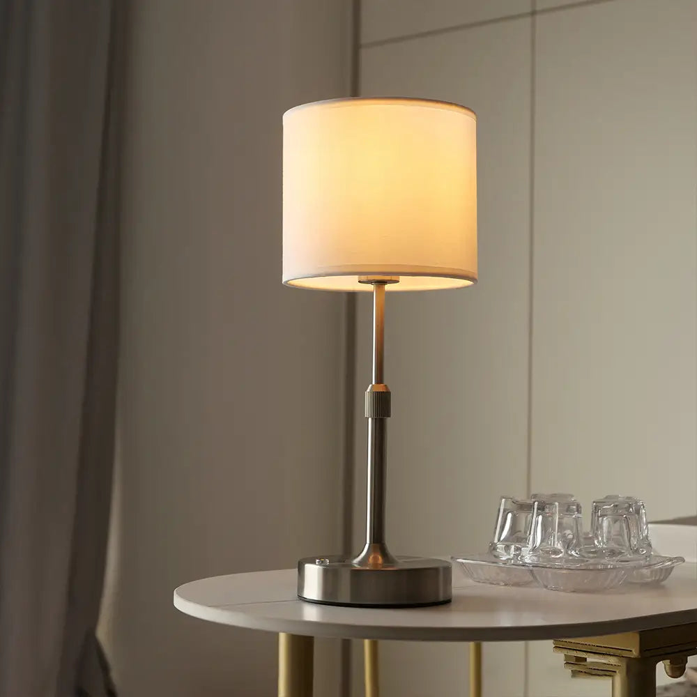 Moderne Touch-Tischlampe mit einem zylindrischen off-weißen Stoffschirm und einer schlichten, gebürsteten Metallbasis, von vorn dargestellt.