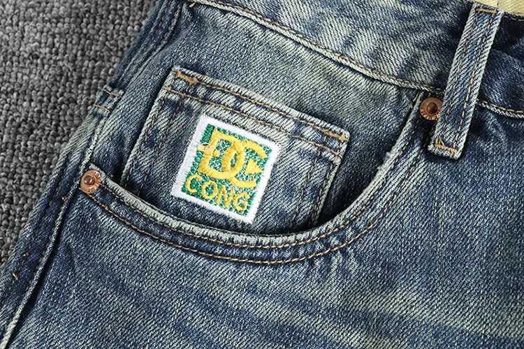 Jeans en denim bleu ample pour hommes présentant un écusson logo DC Comics brodé en blanc, avec un lettrage vert et jaune.