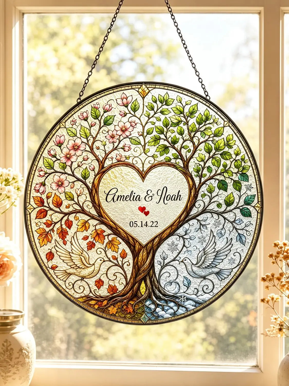 Regalo de boda personalizado de vitrales del árbol de la vida que presenta una decoración colgante de ventana circular con un árbol que muestra el follaje de cuatro estaciones y dos palomas centradas alrededor de un corazón grabado con los nombres Amelia y Noah y la fecha 05.14.22.