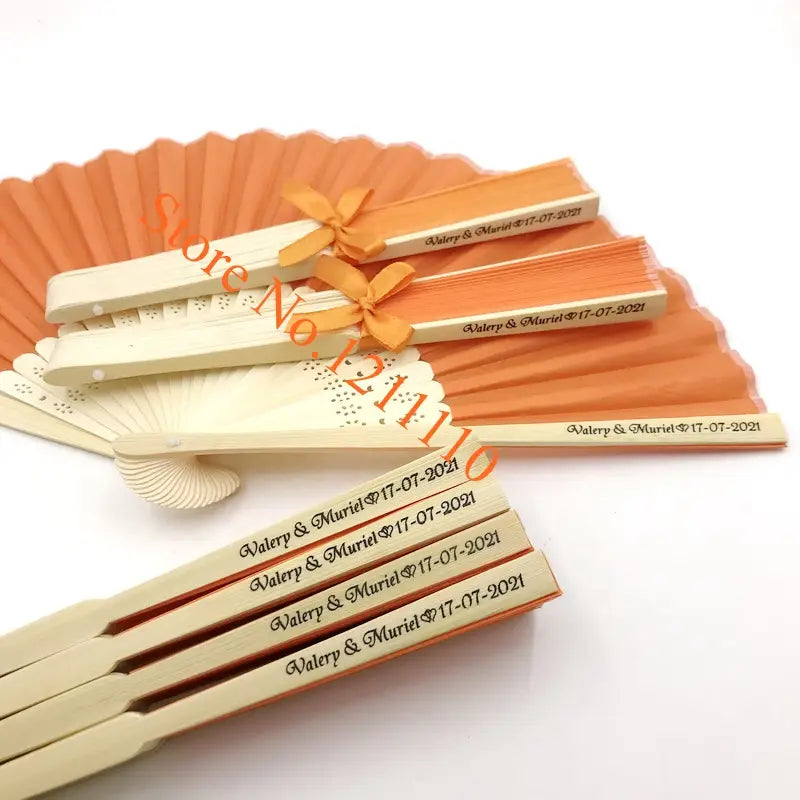 Orangecolored custom wedding hand fans with personalized cream bamboo handles engraved with valery and muriel 17 07 2021 tied together with an orangecolored ribbon made of silk favors.