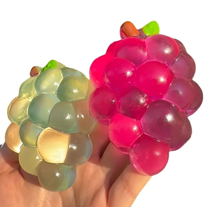 Ein transparenter, glänzender rosa- und grünfarbener Trauben-geformter Stressball, der von einer Hand aus dem Taba-Squishy-Grapefruit-Fidget-Spielzeug Taba-licious Fun Fruit-Kollektion gehalten wird