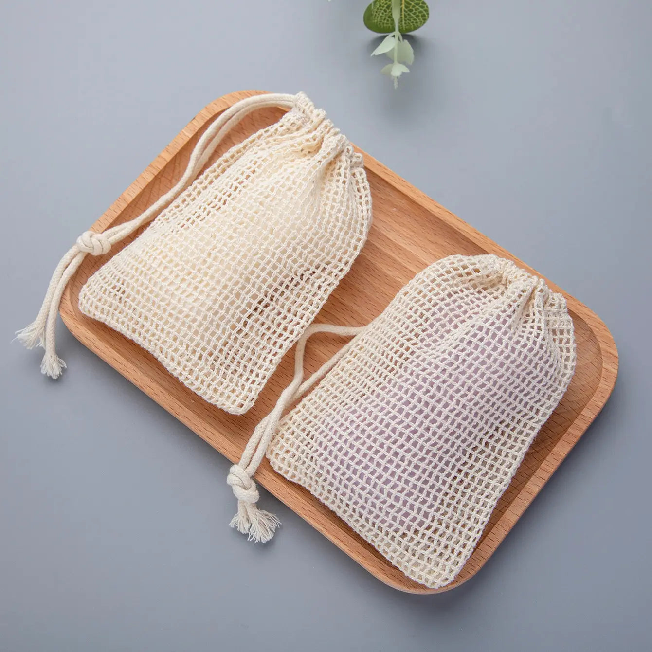 Two off-white cotton soap saver bags made of mesh fabric with drawstrings and frayed ends rest on a wooden tray in a cotton soap saver bag 3 pack.