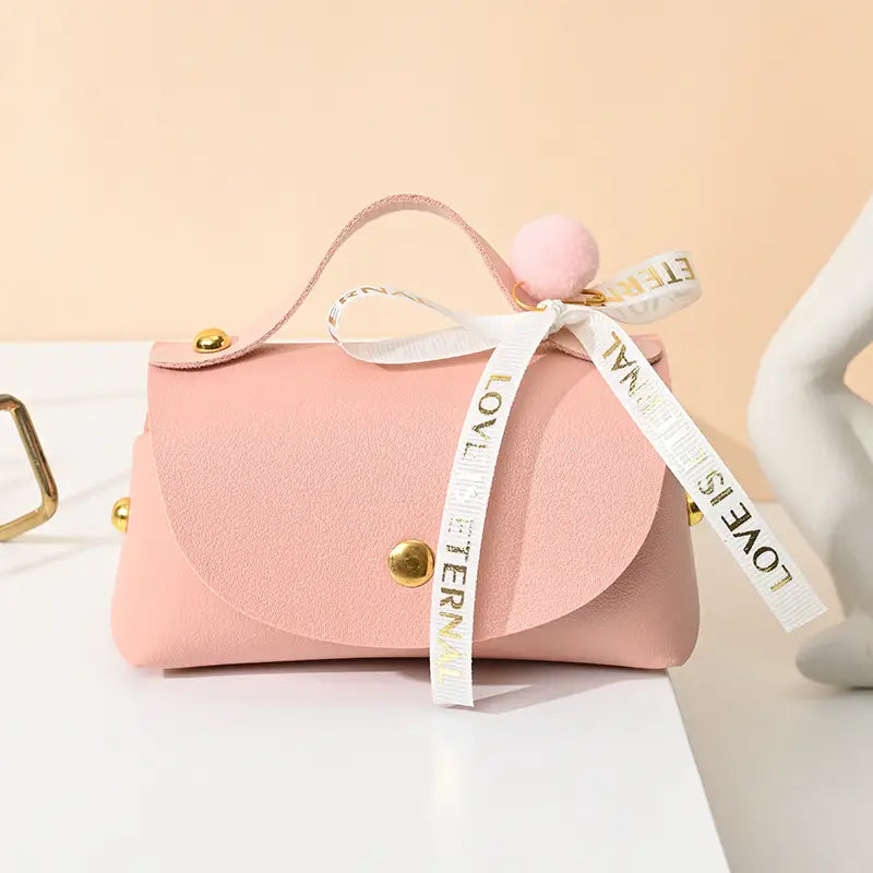 Mini pochette en faux cuir rose tendre avec quincaillerie ton or, un nœud décoratif en ruban blanc portant la mention « l’amour est éternel », et un accent pompon assorti, conçue comme mini sac cadeau de mariage en cuir.